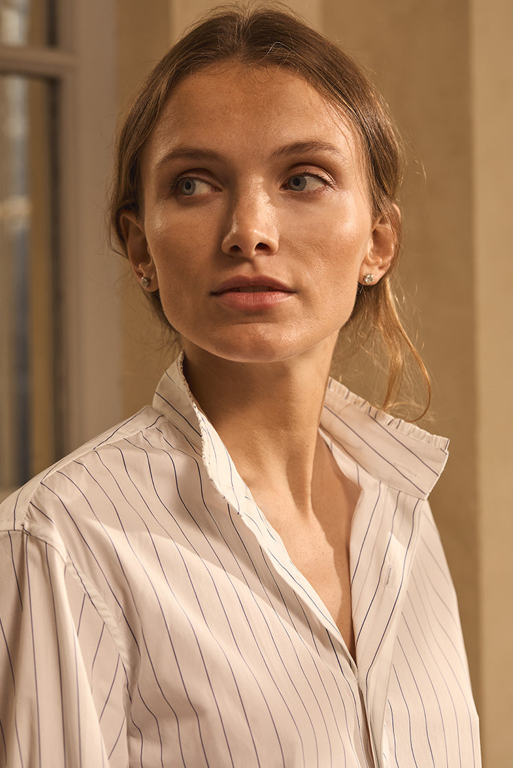 Portrait de femme de trois quart portant une chemise blanche à fines rayures bleu marine, col montant à petits volants.