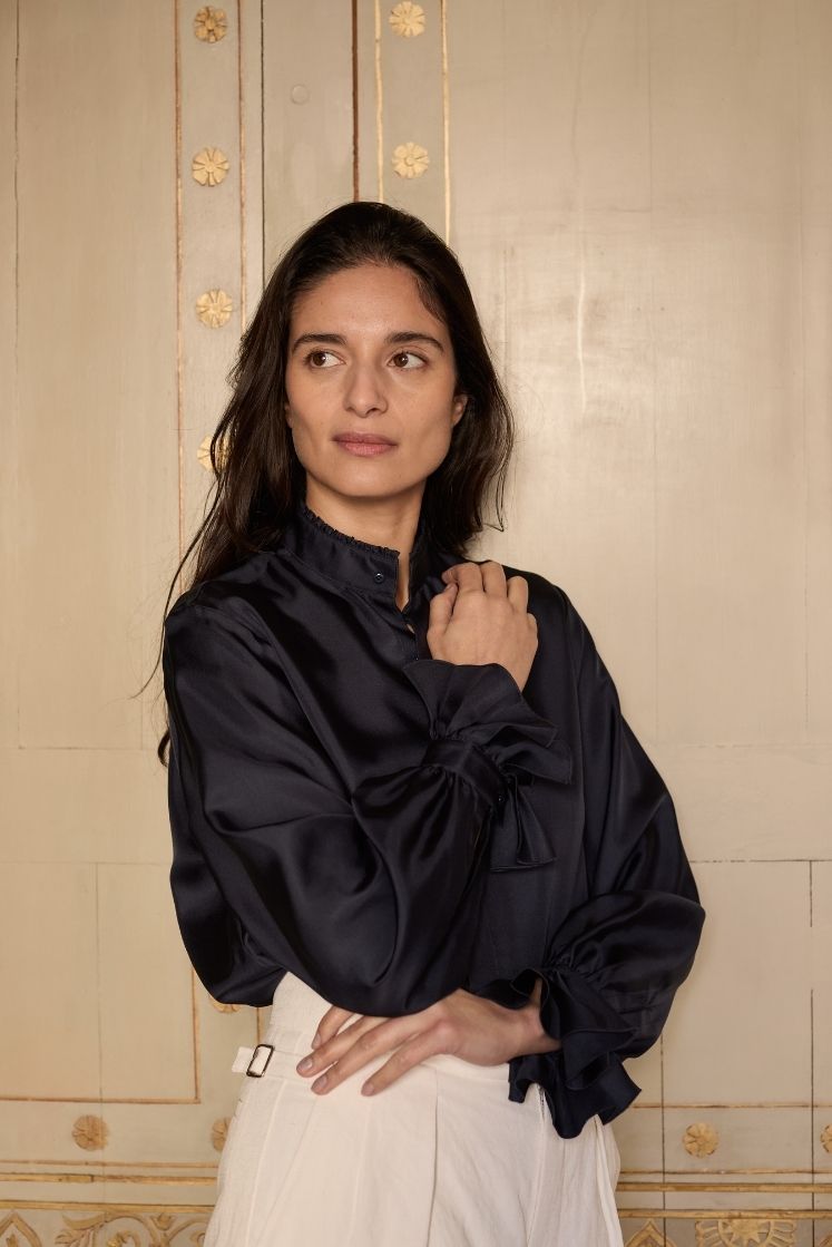 Femme debout dans un décor ancien portant la chemise en soie bleu marine BOUDOIR, avec un col ancien et des poignets à volants, rentrée dans un pantalon en lin blanc.
