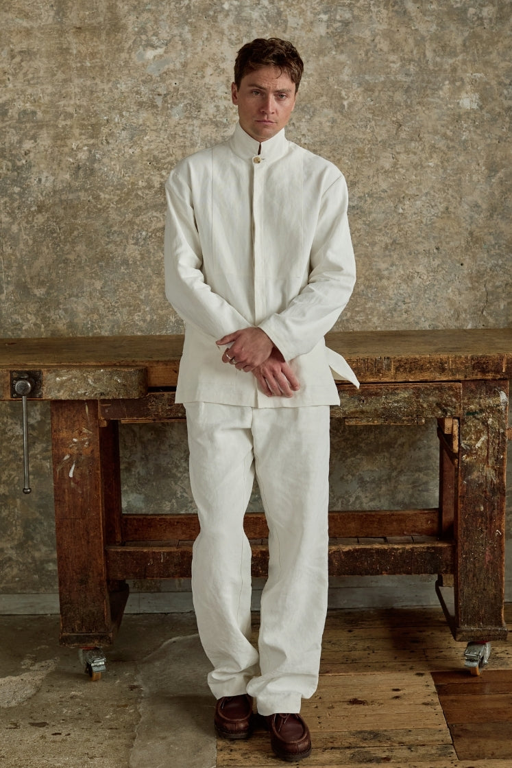 Homme adossé contre une table d'atelier, portant la veste CHEVALET en lin plâtré blanc à col haut, fermée et assortie au pantalon HORIZON en lin plâtré blanc, plan plein pied de face. 
