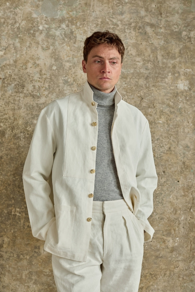 Homme portant la veste CHEVALET en lin plâtré blanc à col haut, ouverte portée par dessus un col roulé gris et assortie au pantalon HORIZON en lin plâtré blanc, plan américain de face. 
