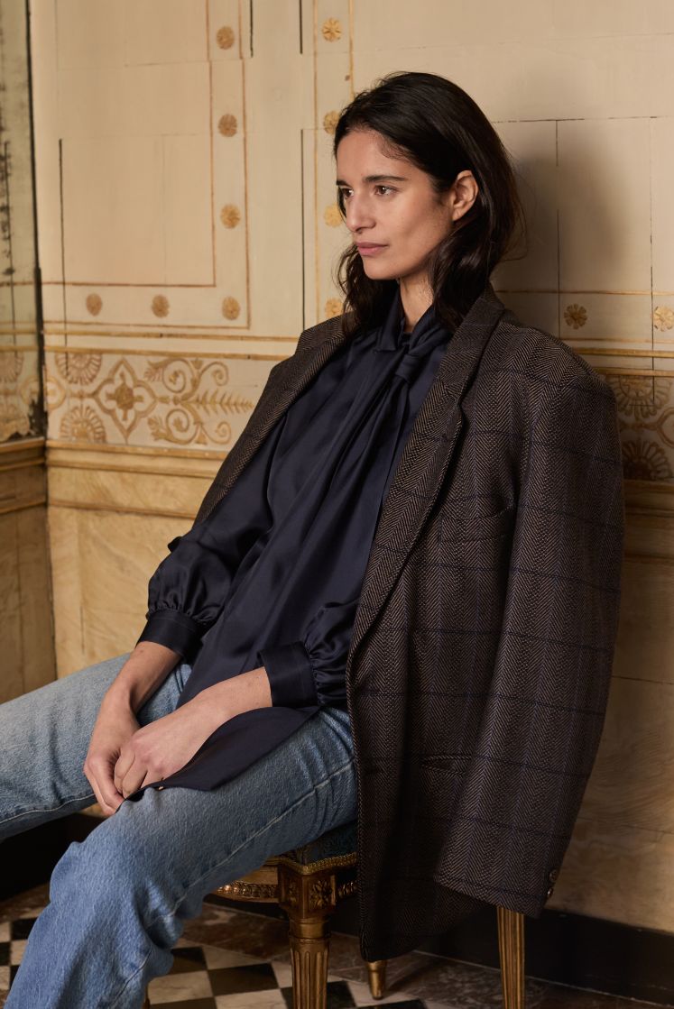 Femme assise dans une chaise, portant la chemise COMTESSE en soie navy noué de façon désinvolte, avec une veste de blazer en laine à carreau et un jean.