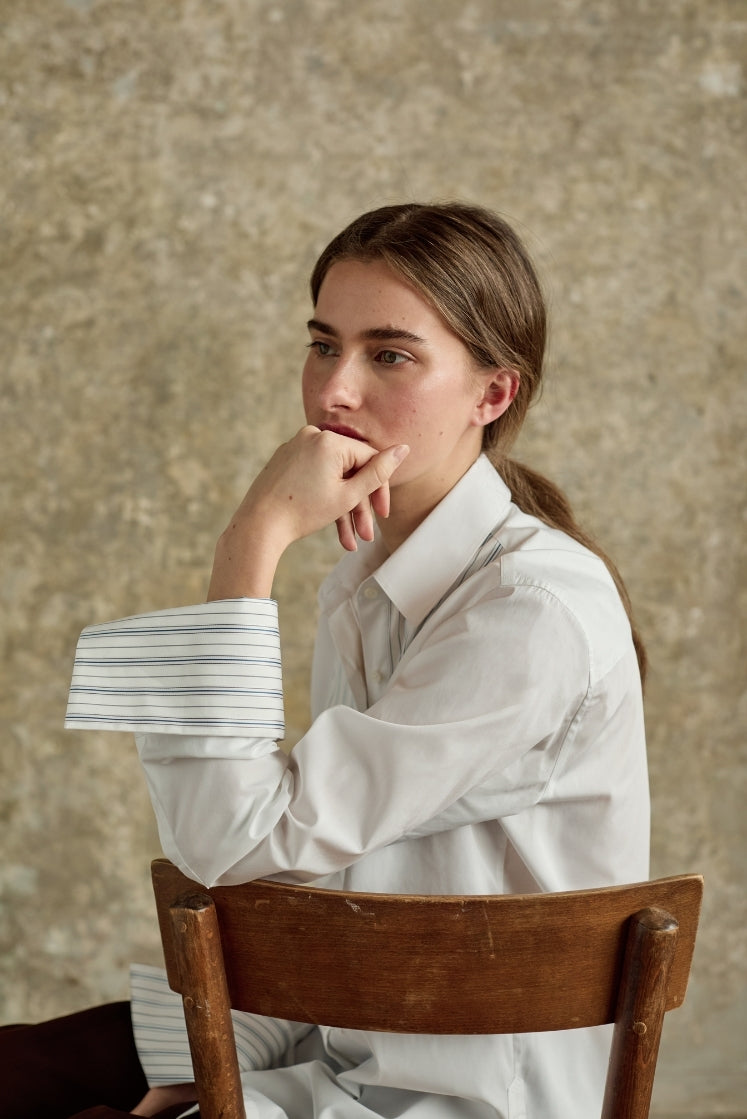 Jeune femme en chemise blanche Bourrienne Paris X, assise pensivement, manches retroussées laissant apparaitre l'intérieur du poignet à rayures, contrastant avec la popeline blanche