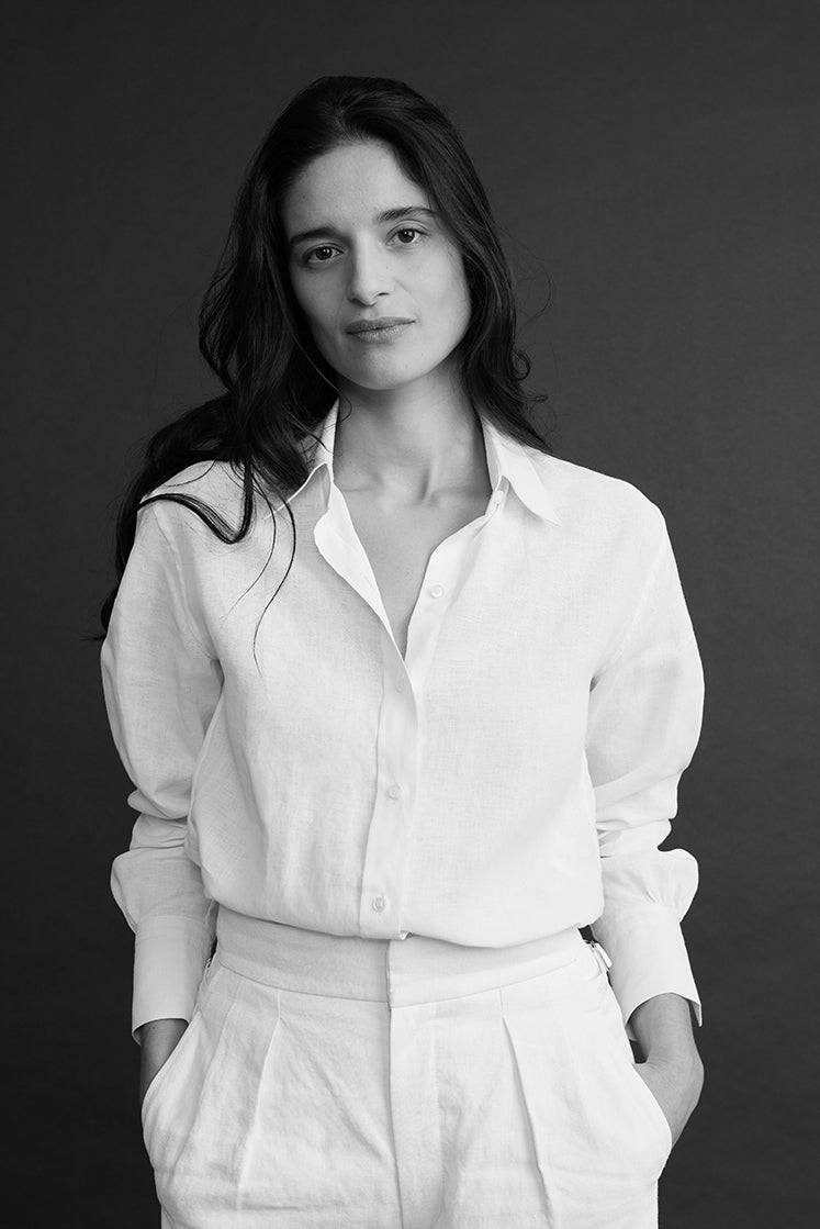 Plan américain d'une femme portant une chemise en lin blanc à col français, légèrement déboutonnée et rentrée dans un pantalon en lin blanc.