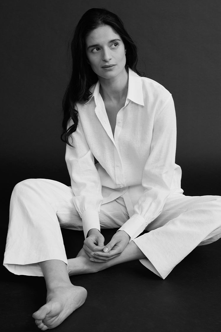 Femme brune assise sur le sol portant une chemise en lin blanc à col français, légèrement déboutonnée, avec un pantalon en lin blanc.