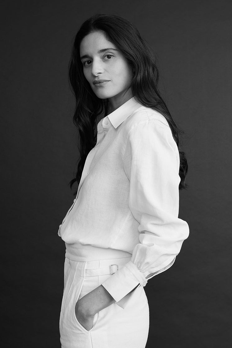 Plan de profil d'une femme brune, posant les mains dans les poches, portant une chemise en lin blanc à col français, rentrée dans un pantalon en lin blanc.