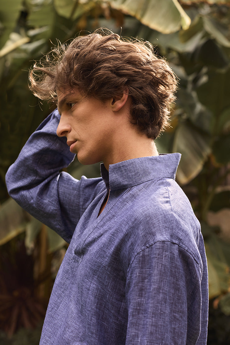 Plan poitrine d'un homme de profil dans un jardin, passant la main dans ses cheveux, vêtu d'une chemise en lin bleu à col haut.