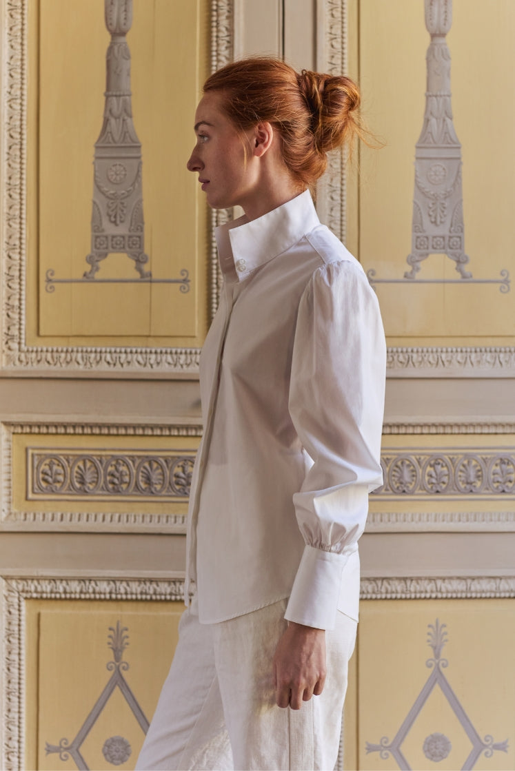 Femme portant la chemise blanche CROISIÈRE à col haut et poignets longs, dans l'Hôtel de Bourrienne, vue de profil. 