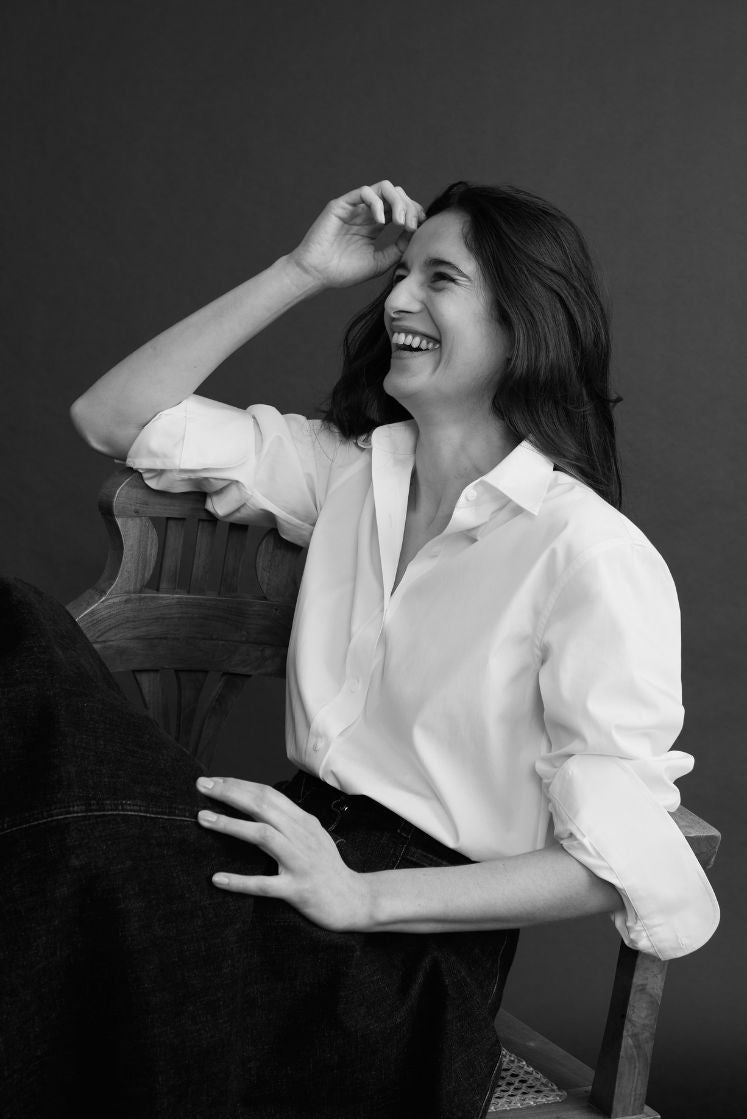 Portrait noir et blanc d’une femme en chemise blanche, col français et poignets classiques, assise sur une chaise en bois, riant avec une attitude naturelle et lumineuse.