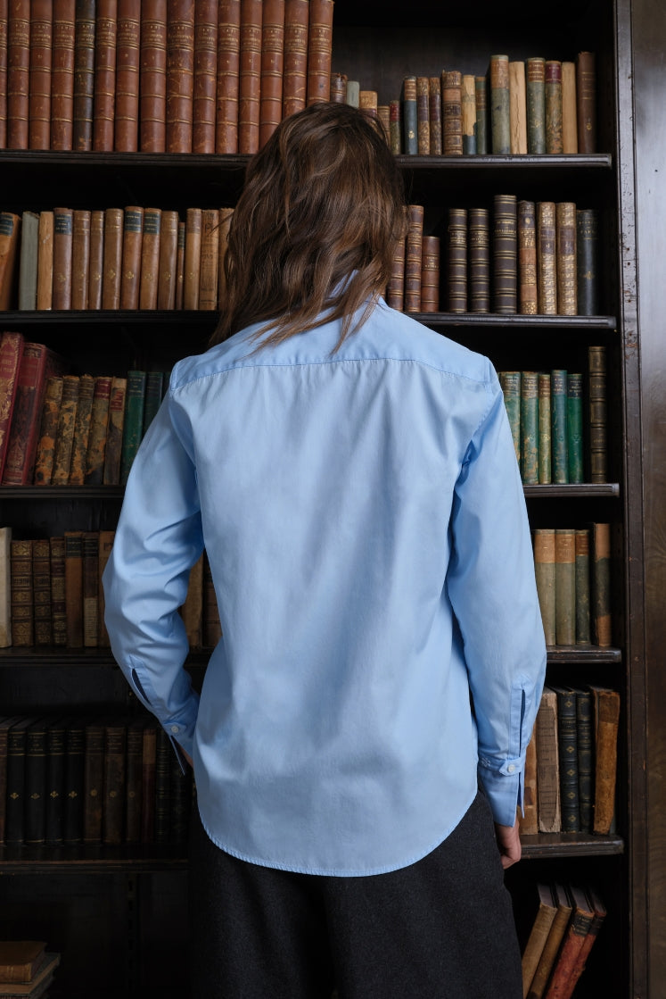 Femme de dos dans une bibliothèque portant une chemise bleu ciel en popeline de coton à la coupe droite.