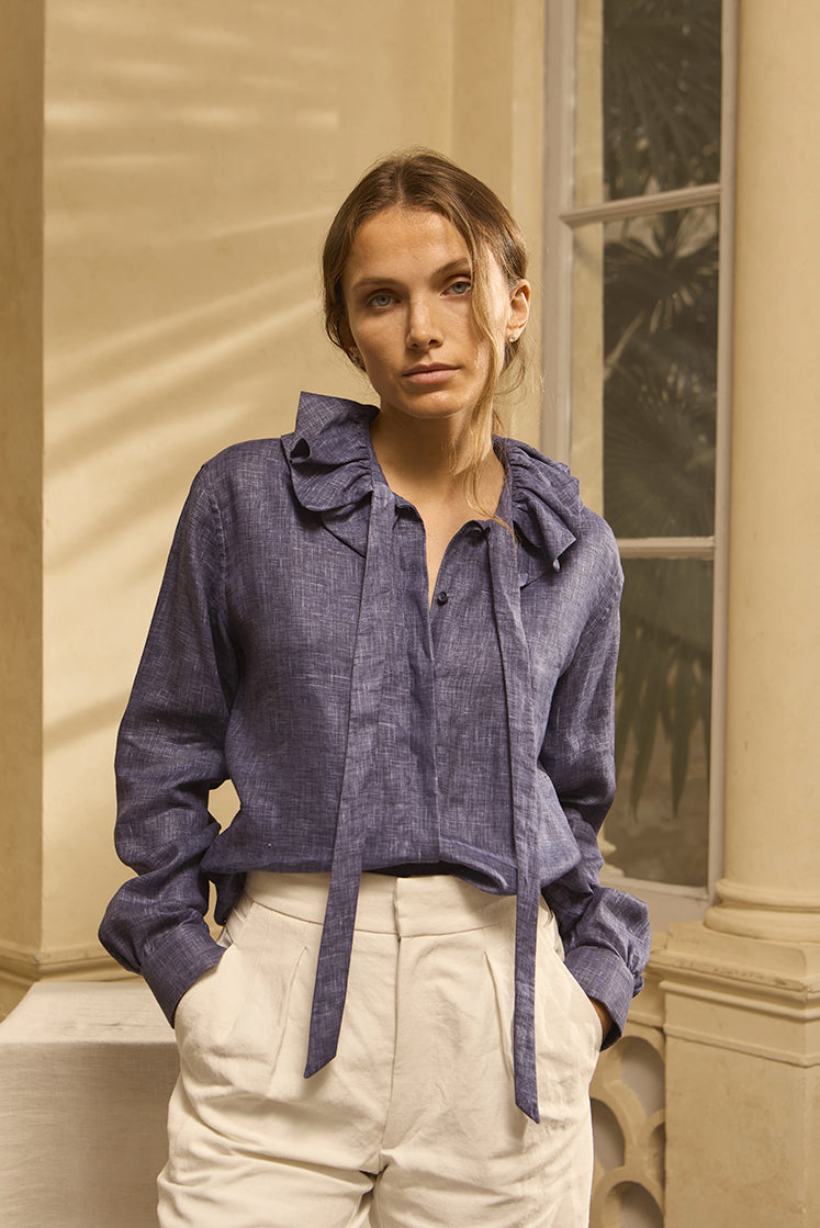 Mannequin portant une chemise en lin leu navy, col à volant, posant les mains dans les poches d'un pantalon en lin blanc.