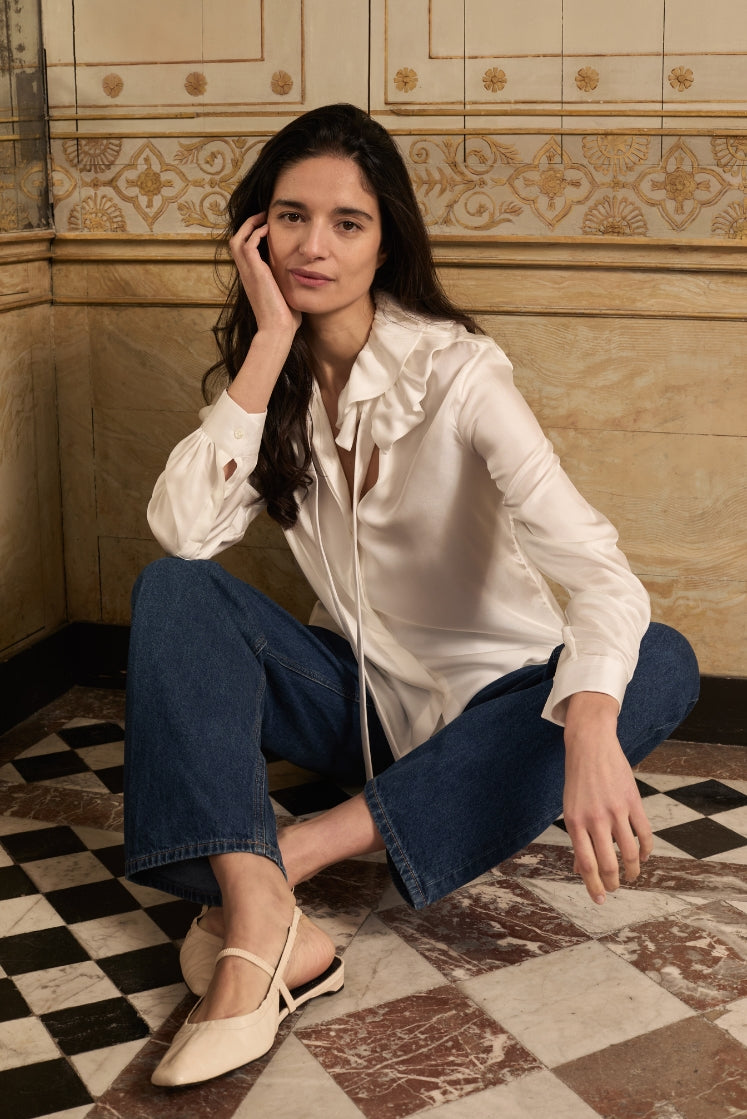 Mannequin assise sur le sol de la salle de bain de l'Hôtel de Bourrienne, portant la chemise PASTEL en soie blanche avec son col à volants, un jean bleu et des ballerines blanches.
