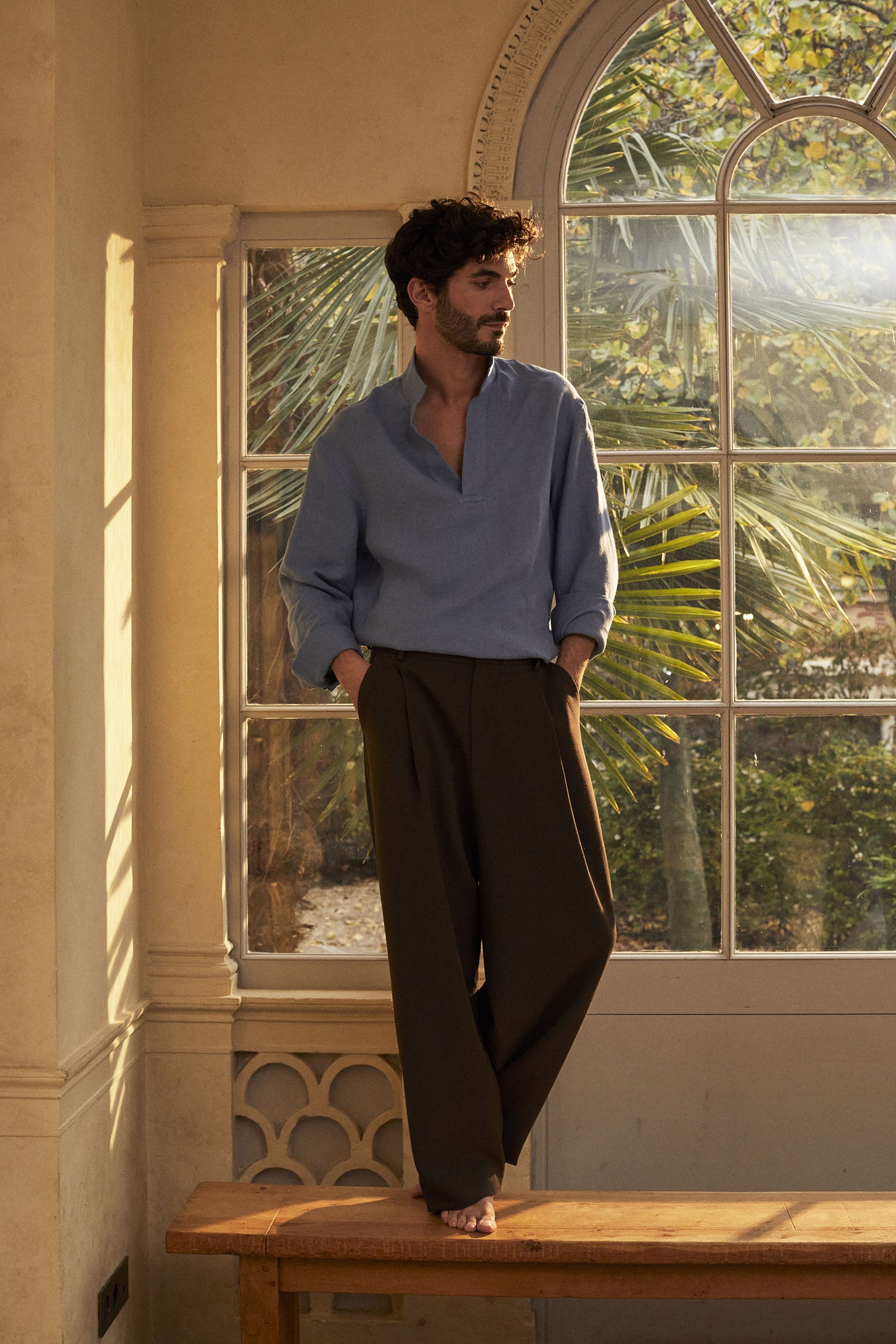 Homme brun portant la chemise Bourrienne Paris X en lin bleu, posant pieds nus sur un banc en bois, baigné par la lumière dorée d'une verrière avec la nature en arrière-plan.