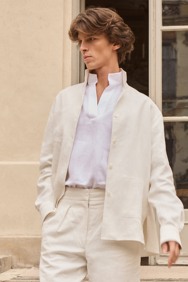 Homme portant un look blanc avec une este et un pantalon en lin épais, et une chemise à col haut en lin fin, une main dans sa poche, regardant l'horizon.