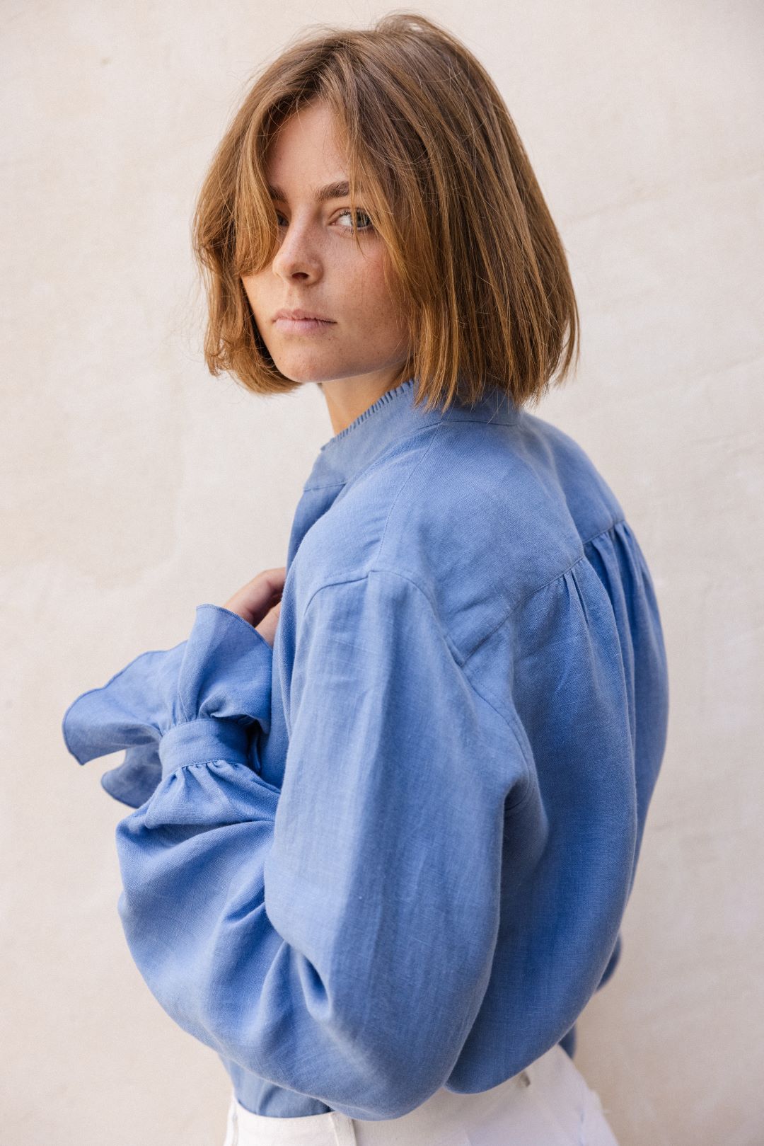 Photo en plan américain, une femme blonde aux cheveux carrés est de profil et ses yeux fixent l'objectif. Elle porte une chemise BOUDOIR en Lin Bleu confectionnée dans un lin doux et léger, teinté d’un bleu ciel délicat, avec un col officier délicatement bordé de petits volants pour une touche raffinée et ses manches légèrement bouffantes agrémentées de volants qui subliment la silhouette.